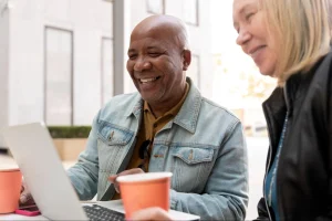 Close-up de um homem e uma mulher sorrindo enquanto olham para a tela de um laptop. Eles estão em um ambiente iluminado, possivelmente uma cafeteria, com copos de café em primeiro plano. A expressão deles é de satisfação e entusiasmo.
