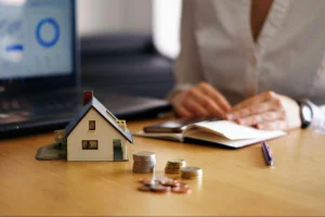Em primeiro plano, uma casa em miniatura e pilhas de moedas estão organizadas sobre uma mesa de madeira. No fundo desfocado, uma pessoa sentada segura uma caneta sobre um caderno aberto, com um laptop mostrando gráficos ao lado. A imagem evoca o conceito de planejamento financeiro e investimento imobiliário.