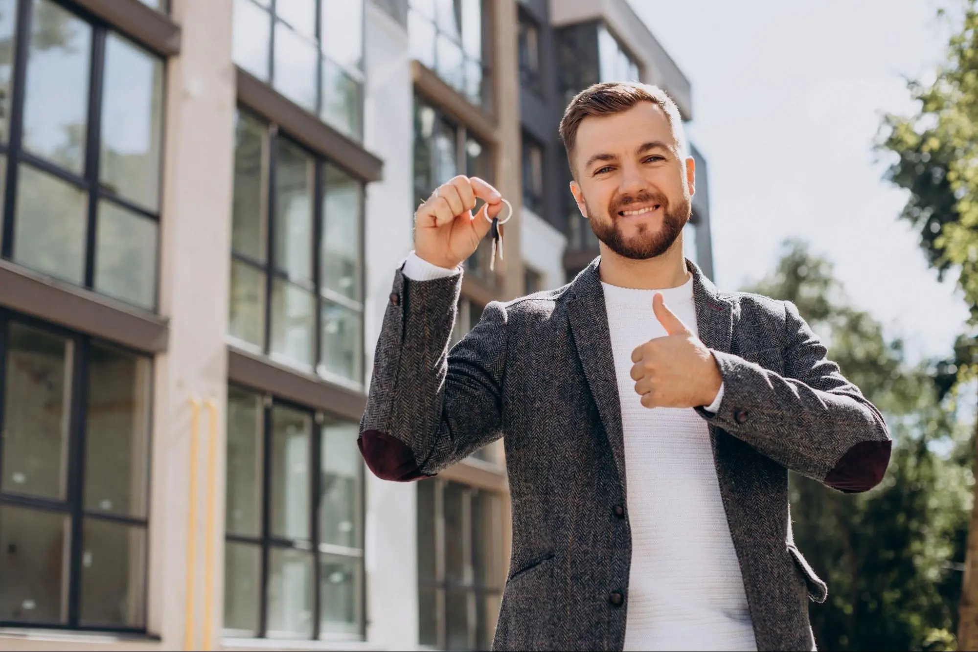 Homem jovem sorridente, vestindo blazer cinza e camiseta branca, segura as chaves de um novo imóvel e faz um sinal de positivo com o polegar. Ao fundo, a fachada moderna de um prédio residencial com grandes janelas de vidro reflete a luz do dia.