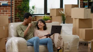 Casal interracial sorridente usa notebook em sofá coberto de plástico, cercado por caixas de papelão. Eles parecem planejar a decoração ou compras online para o novo apartamento durante a mudança.