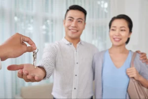 Em primeiro plano, a mão de um corretor entrega chaves para a mão estendida de um homem. Ao fundo, um jovem casal asiático sorri, desfocado, celebrando a conquista do novo imóvel.