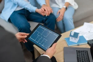 Corretor segurando um tablet com planta de apartamento exibida na tela, em frente a um casal sentado no sofá de mãos dadas, com maquete de casa, notebook e documentos sobre a mesa.