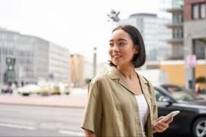 Mulher jovem caminha pela rua do centro e consulta o celular para usar mapa, em meio a prédios e movimento urbano.