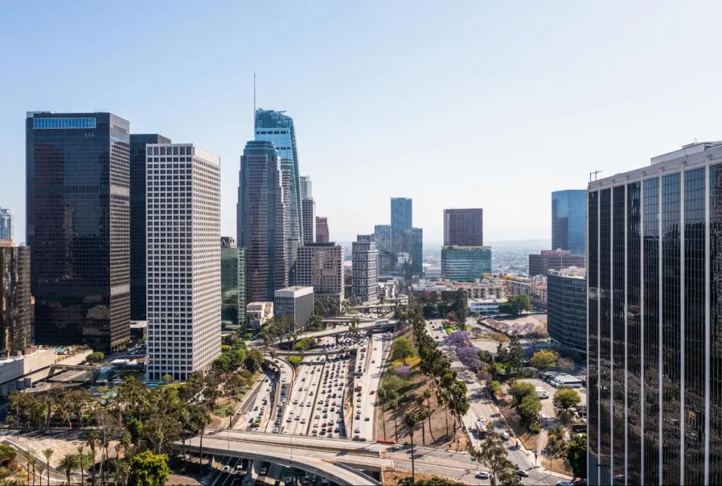 Vista aérea de centro urbano com grandes avenidas, arranha-céus, tráfego intenso e áreas de infraestrutura entre os edifícios.