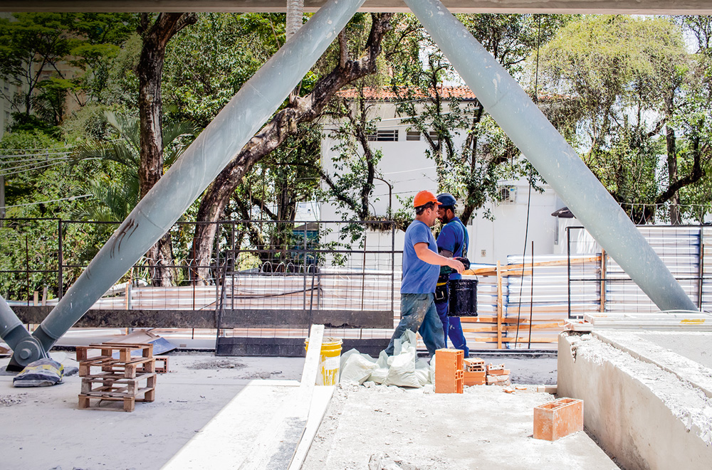 Imagem mostra dois trabalhadores de capacete e camiseta azul em canteiro de obras com tijolos e cimento fresco.
