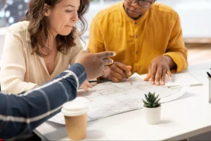 Grupo de pessoas em mesa de reunião analisando planta de apartamento impressa, fazendo anotações e discutindo detalhes técnicos do imóvel.