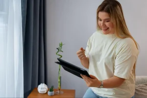 Mulher sentada em quarto iluminado segurando tablet, com uma mesa lateral decorada com plantas ao fundo.
