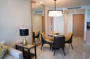 Sala de jantar moderna, com mesa redonda posta para quatro pessoas, cadeiras em tons de preto e amarelo, lustre pendente, espelho grande na parede e decoração elegante em tons neutros.