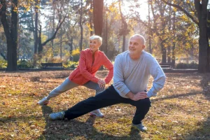 Casal idoso sorrindo faz exercício de alongamento em um parque arborizado, em um dia ensolarado de outono.