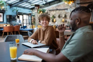 Dois amigos conversam em uma cafeteria moderna, sentados à mesa com bebidas e notebook em um ambiente acolhedor.