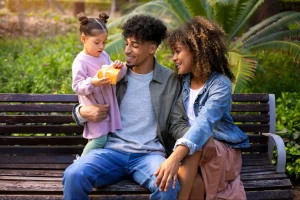 Família de três pessoas aproveita um momento ao ar livre em um parque, com criança no colo e clima de convivência em meio à natureza.