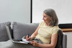 Uma mulher de meia-idade vista de perfil, está confortavelmente sentada em um sofá cinza utilizando um notebook. Suas pernas estão cobertas por uma manta com padrão zigue-zague cinza e branco.