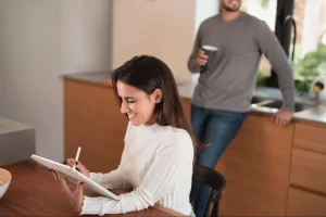 Uma mulher jovem de perfil, sorridente e com cabelos longos e escuros, está sentada à mesa de madeira utilizando uma caneta digital em um tablet. Ela veste uma blusa branca de gola alta. Ao fundo, em um plano mais afastado e ligeiramente desfocado, um homem de camiseta cinza de manga longa e calça jeans está de pé na cozinha americana, encostado na bancada de madeira enquanto segura uma caneca.