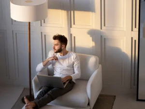 Homem sentado em poltrona branca com apoio para os pés, usando notebook. Ele está sendo iluminado por luz natural suave em ambiente moderno e minimalista, com painel de parede quadriculado e luminária de chão.