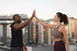 Homem e mulher treinando juntos ao ar livre, dando um toque de mão em gesto de comemoração, com prédios ao fundo no entardecer.