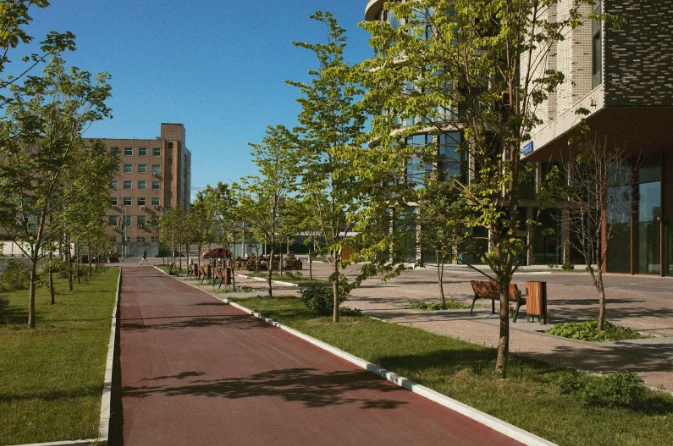 Caminho arborizado com bancos ao lado de edifícios modernos em uma área urbana, destacando a integração entre natureza e cidade.