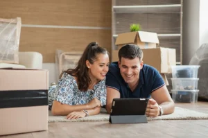 Casal jovem deitado no chão, cercado por caixas de mudança, usando um tablet para planejar detalhes do novo apartamento e avaliar localização estratégica.