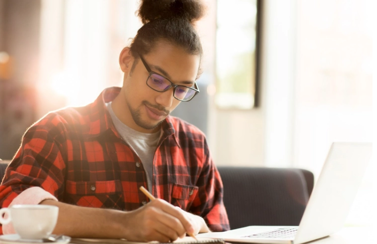 Homem com óculos e camisa xadrez trabalhando em um caderno ao lado de um notebook, sentado à mesa em ambiente iluminado.