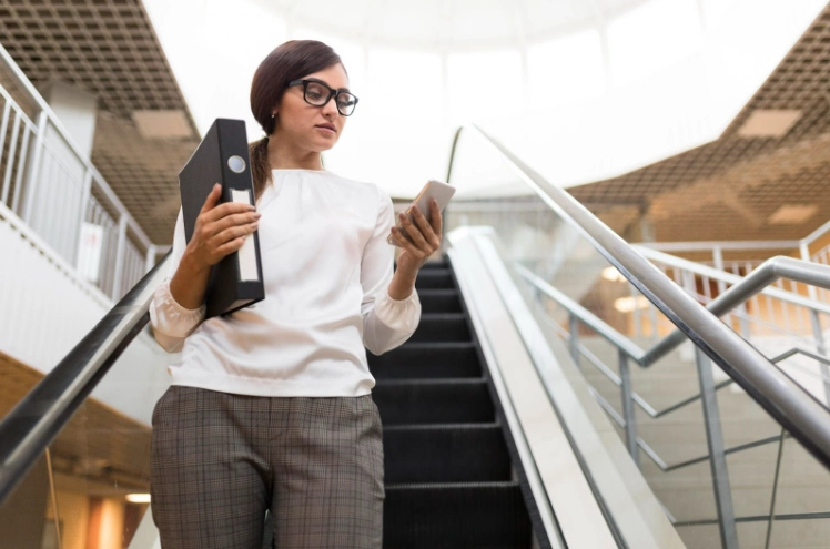 Mulher usando óculos, segurando um fichário e olhando o smartphone enquanto desce uma escada em ambiente corporativo moderno, com iluminação natural ao fundo.