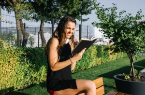 Mulher sentada em banco de parque lendo um livro, cercada por vegetação e árvores em meio ao centro urbano.