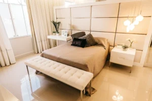 Quarto moderno e bem iluminado com uma cama de casal, decoração em tons neutros, plantas, luminárias pendentes e móveis funcionais, destacando o aproveitamento inteligente do espaço.
