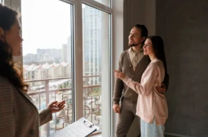 Casal jovem observa a paisagem urbana através da janela de um apartamento novo, acompanhado por uma agente imobiliária que apresenta o imóvel.