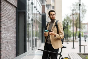 Homem jovem com estilo moderno, segurando bicicleta, café para viagem e celular, parado na calçada de uma área urbana com prédios ao fundo, vestindo blazer bege, mochila preta e óculos.
