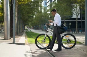 Homem de negócios empurrando bicicleta e usando celular em rua arborizada com trilhos, simbolizando o uso de alternativas sustentáveis para mobilidade urbana.