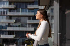 Mulher de cabelo longo em varanda de edifício moderno, observando as fachadas envidraçadas de outros apartamentos, sob luz natural.
