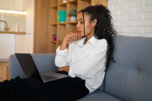 Mulher de cabelos cacheados sentada no sofá, vestindo roupa clara, com notebook no colo. Ela aparenta estar pensativa em ambiente de sala moderna integrada à cozinha.