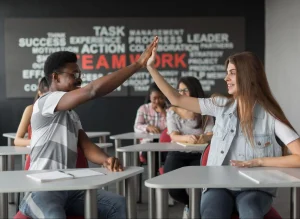 Dois estudantes em sala de aula comemoram, sorrindo e sentados em carteiras, com colegas ao fundo e a palavra “TEAMWORK” destacada na parede.