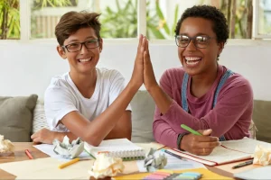 Dois jovens, sorrindo e usando óculos, dão “high five” enquanto estudam juntos em uma mesa coberta de materiais escolares e papéis, demonstrando cooperação e entusiasmo.