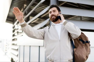 Homem sorridente com barba, falando ao celular e acenando, usando jaqueta clara e mochila, em área externa de aeroporto, transmitindo praticidade e boa disposição.
