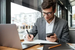 Jovem empresário de óculos, sorridente, sentado em uma cafeteria, faz anotações em um caderno enquanto usa o smartphone e trabalha com notebook na mesa.