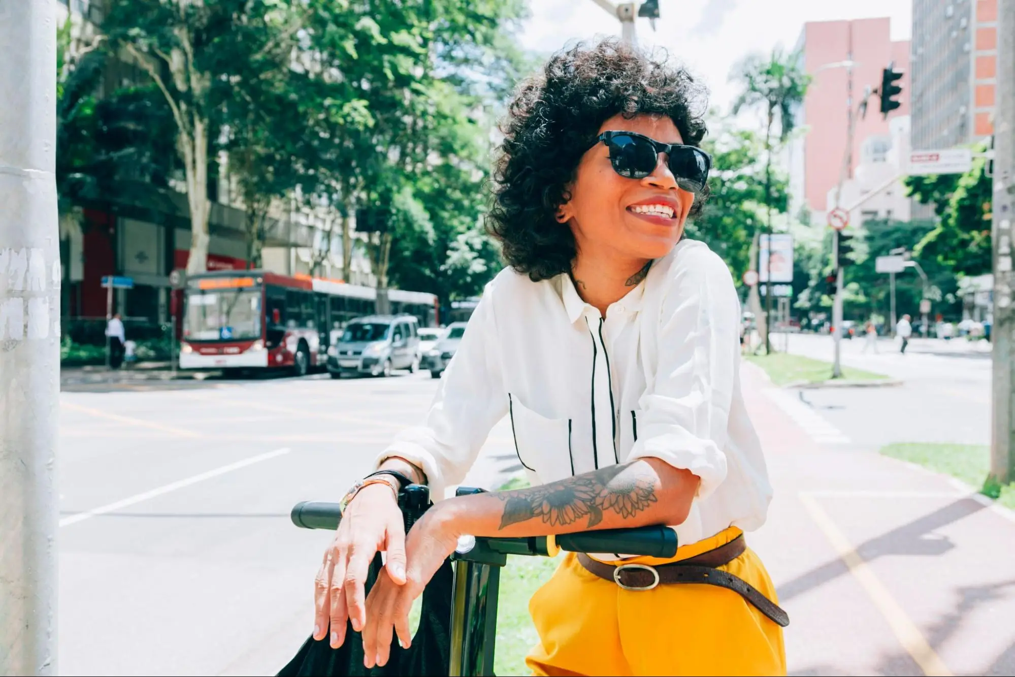 Mulher sorridente com óculos escuros, camisa branca e calça amarela, segura uma scooter elétrica em uma avenida arborizada de cidade grande.
