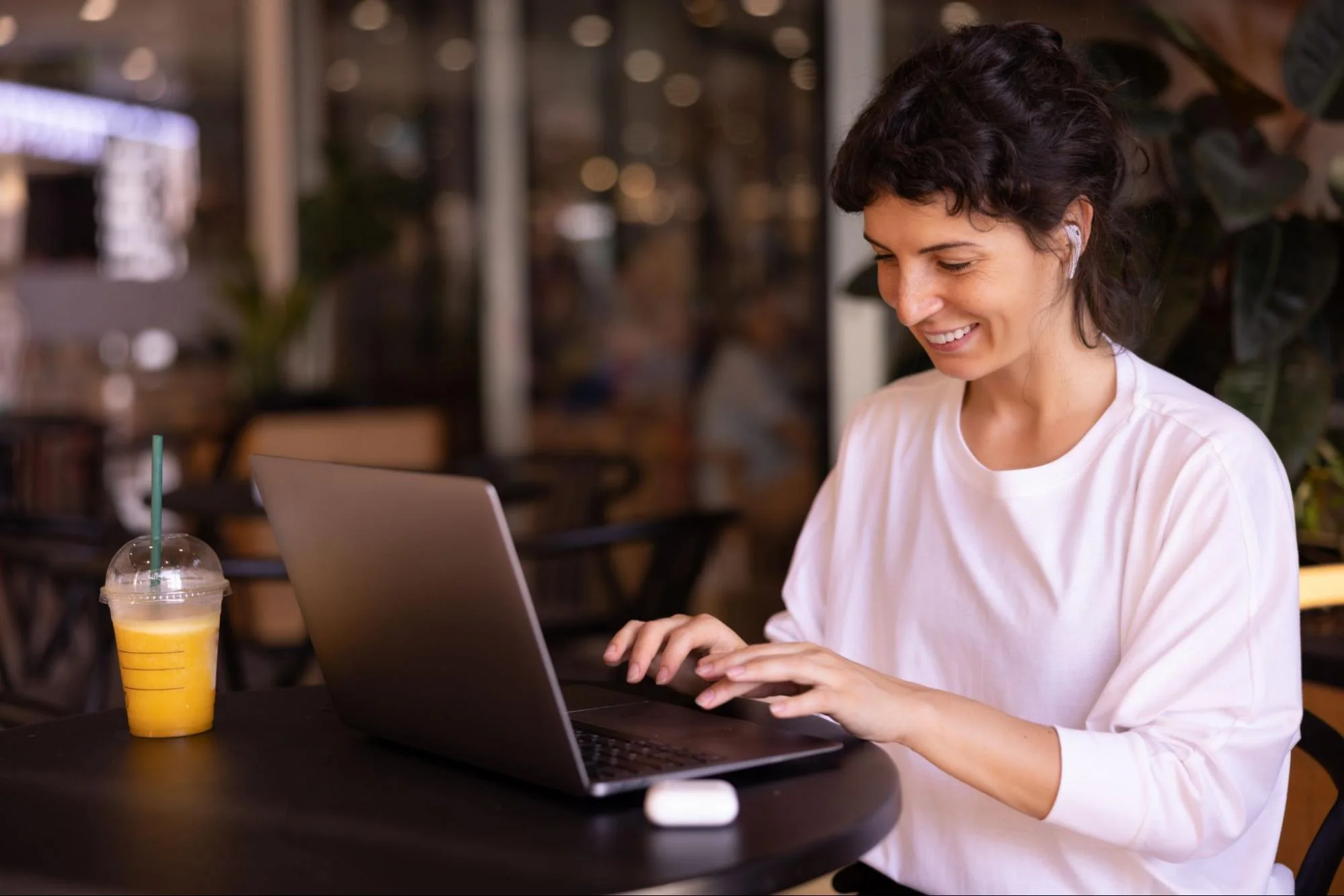 Mulher trabalhando em um laptop em cafeteria moderna, acompanhada de um suco, em clima descontraído no bairro urbano.