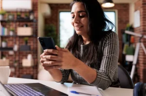 Mulher sentada à mesa, usando o celular e com notebook aberto ao lado, transmitindo praticidade em ambiente residencial.