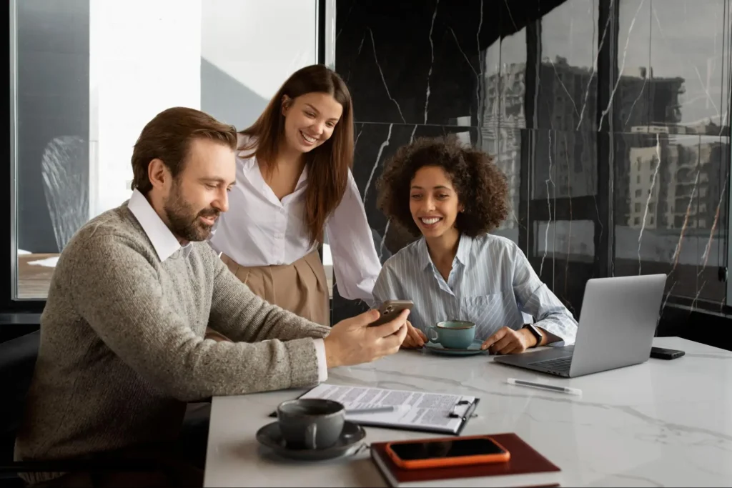 Três colegas de trabalho sorriem enquanto conversam em uma mesa de escritório moderna, usando notebook e celular, em ambiente com grandes janelas e vista urbana ao fundo.