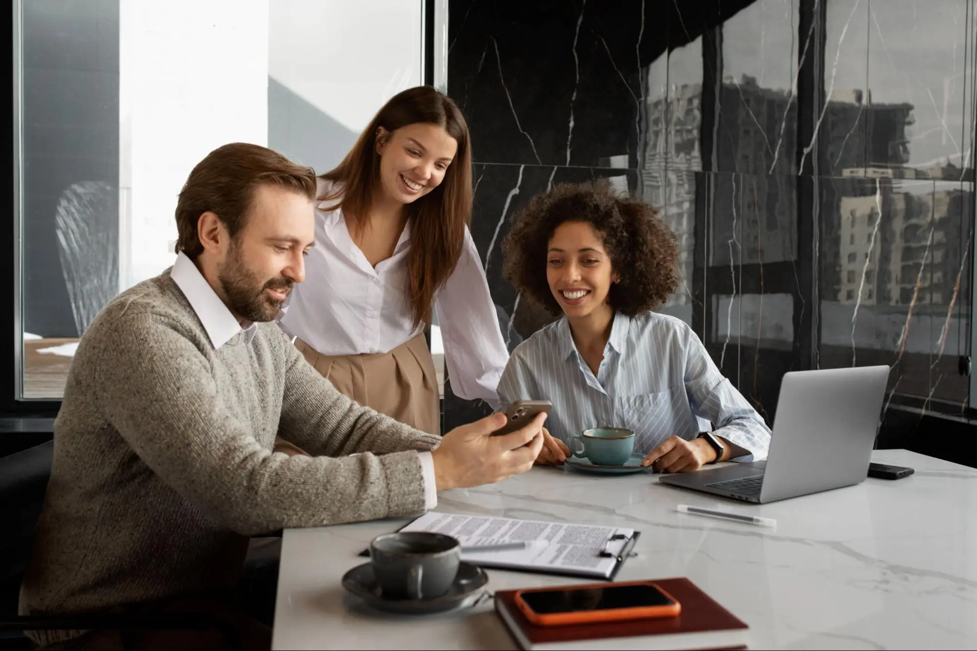 Três colegas de trabalho sorriem enquanto conversam em uma mesa de escritório moderna, usando notebook e celular, em ambiente com grandes janelas e vista urbana ao fundo.