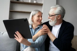 Casal de idosos sentados no sofá, sorrindo e usando juntos um tablet, transmitindo tranquilidade e conexão em ambiente moderno e acolhedor.