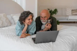 Casal deitado sobre a cama, olhando para a tela de um notebook enquanto um deles segura um gato no colo em um quarto aconchegante.