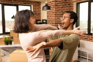 Casal jovem em ambiente interno com caixas de mudança ao fundo, abrindo os braços e sorrindo um para o outro em clima de comemoração dentro da nova casa.