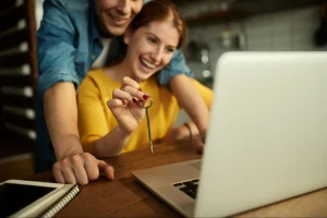 Casal jovem sentado à mesa em frente ao notebook, sorrindo enquanto a mulher mostra uma chave de imóvel em destaque para a câmera.
