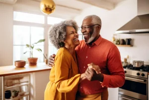 Casal maduro dançando abraçado na cozinha de um apartamento iluminado, com janelas amplas ao lado