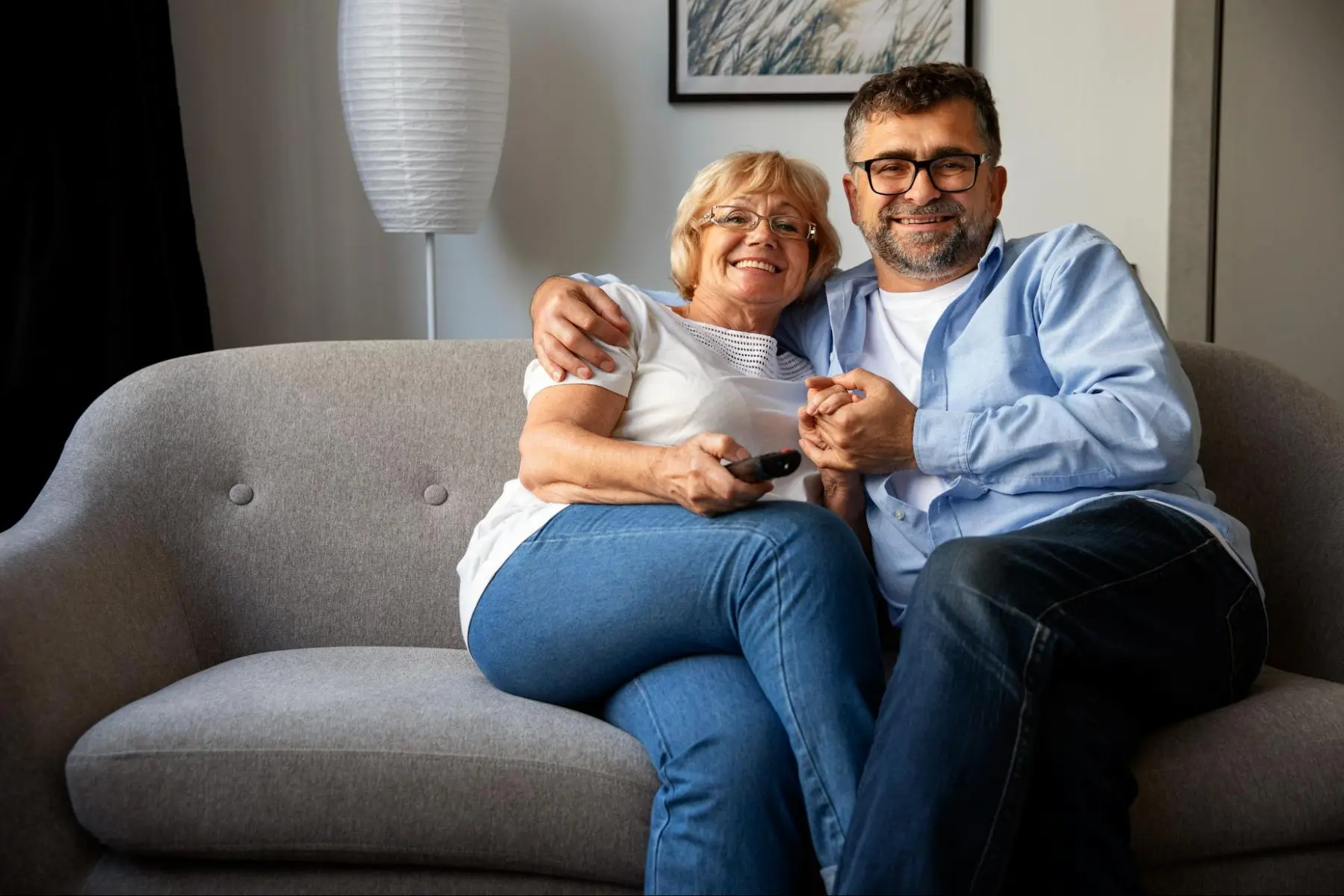 Casal maduro sentado junto em um sofá cinza, abraçado e assistindo televisão na sala de estar.