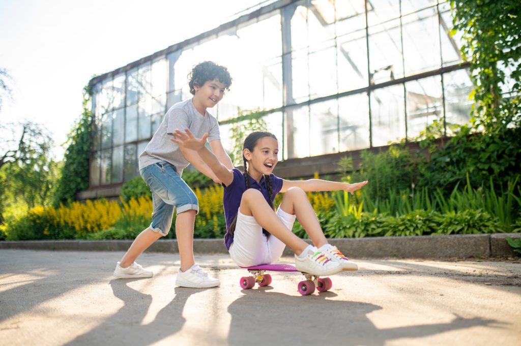 Duas crianças brincando ao ar livre. Uma está sentada em um skate enquanto a outra empurra, em uma área com vegetação e estufa ao fundo em dia ensolarado.