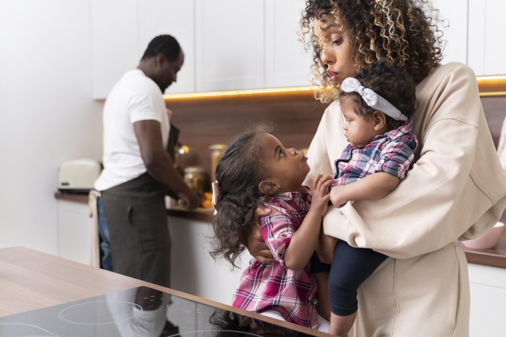 Mãe segurando um bebê no colo e abraçando a filha mais velha na cozinha, enquanto o pai prepara algo no balcão ao fundo.