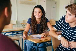 Adolescente sentada em carteira escolar, com caderno e lápis sobre a mesa, conversando com colegas em uma sala de aula.