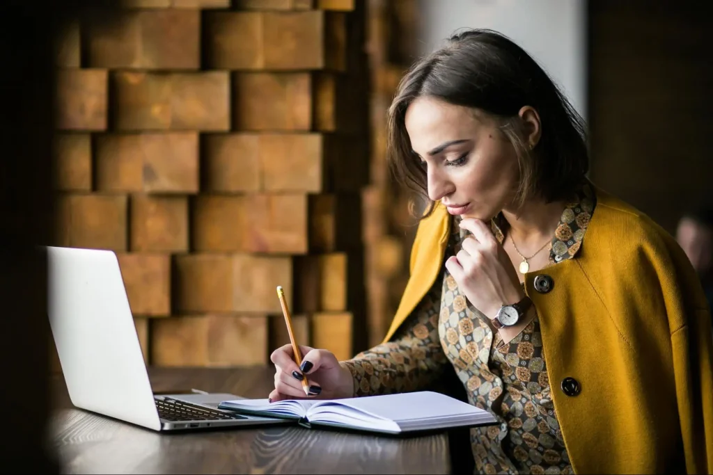 Mulher jovem sentada em um café trabalhando em um notebook, escrevendo em um caderno com lápis e usando um casaco mostarda sobre uma camisa estampada, com parede de madeira ao fundo.