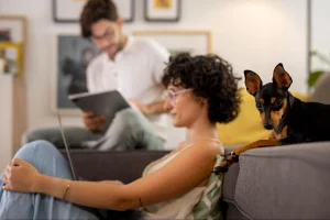 Mulher sentada no chão usando notebook enquanto um cachorro pequeno está deitado no sofá, com um homem ao fundo usando um tablet.